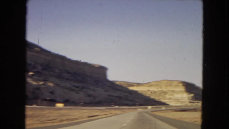 A passenger-view highway shot shows the road ahead cutting through layered desert terrain, with no people visible in the frame.