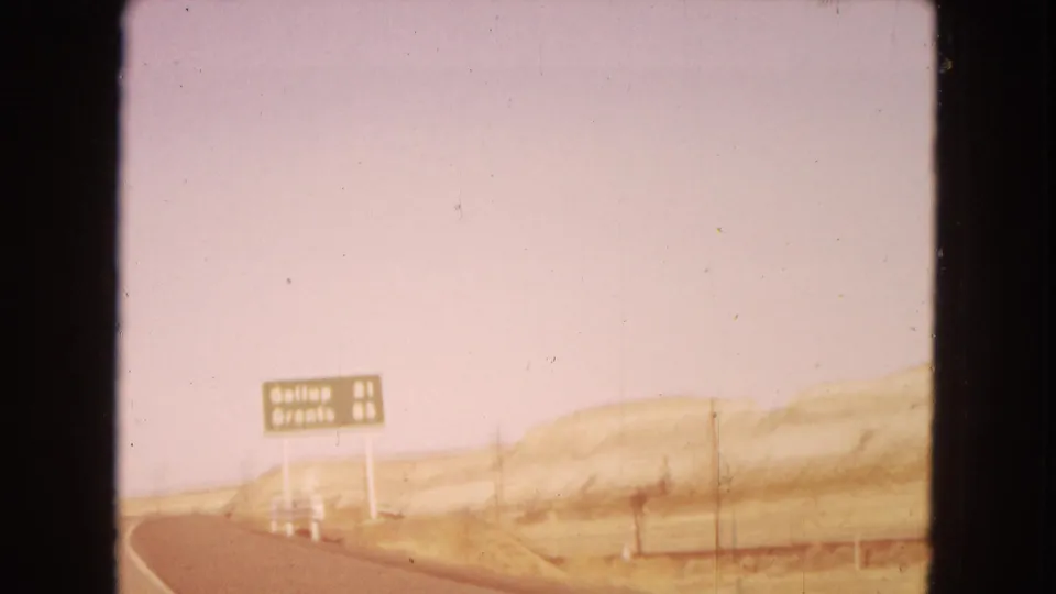 A roadside direction sign for Gallup and Grants appears beside the highway in the Southwestern desert, seen through faded 8mm color.