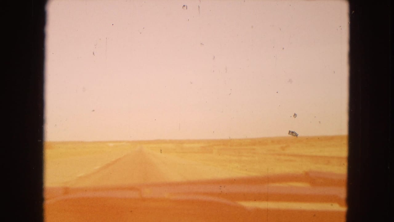 1967 Arizona home-movie view from inside a moving vehicle, looking through a dusty windshield toward a straight desert road and flat horizon.