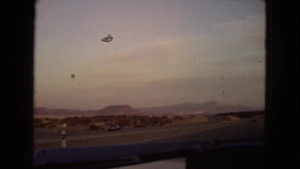 Arizona desert roadside view with low foreground, distant mesa-like formations, open sky, and dark film-edge framing from a moving home-movie perspective.