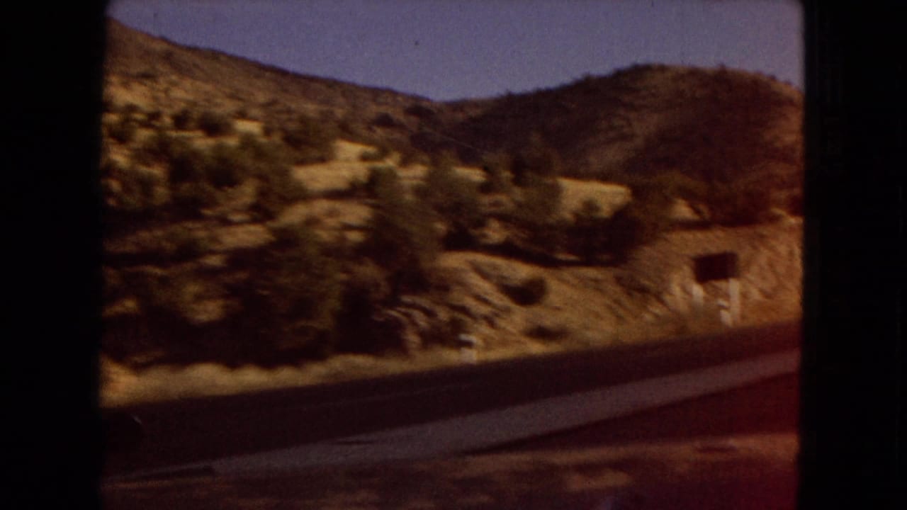 1967 Arizona road footage showing dry hills, sparse shrubs, a roadside barrier, and a small sign passing outside the car window.