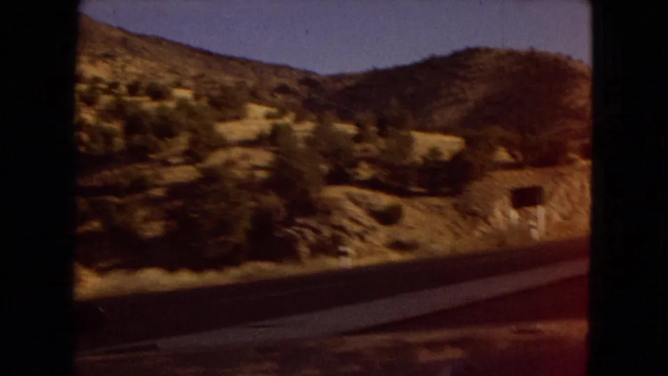 1967 Arizona road footage showing dry hills, sparse shrubs, a roadside barrier, and a small sign passing outside the car window.
