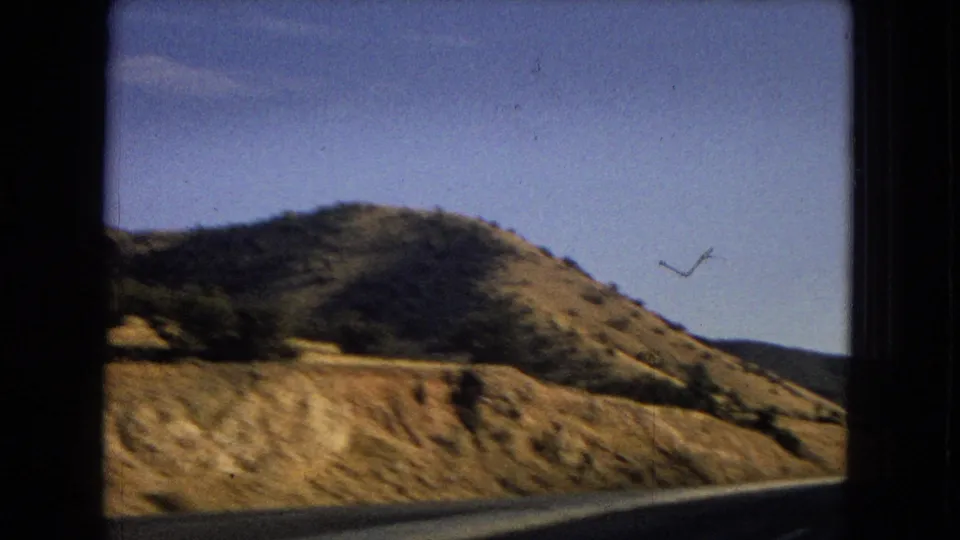 1967 8mm frame of an Arizona roadside curve below dry cut slopes, scrub vegetation, and low hills under a blue sky.