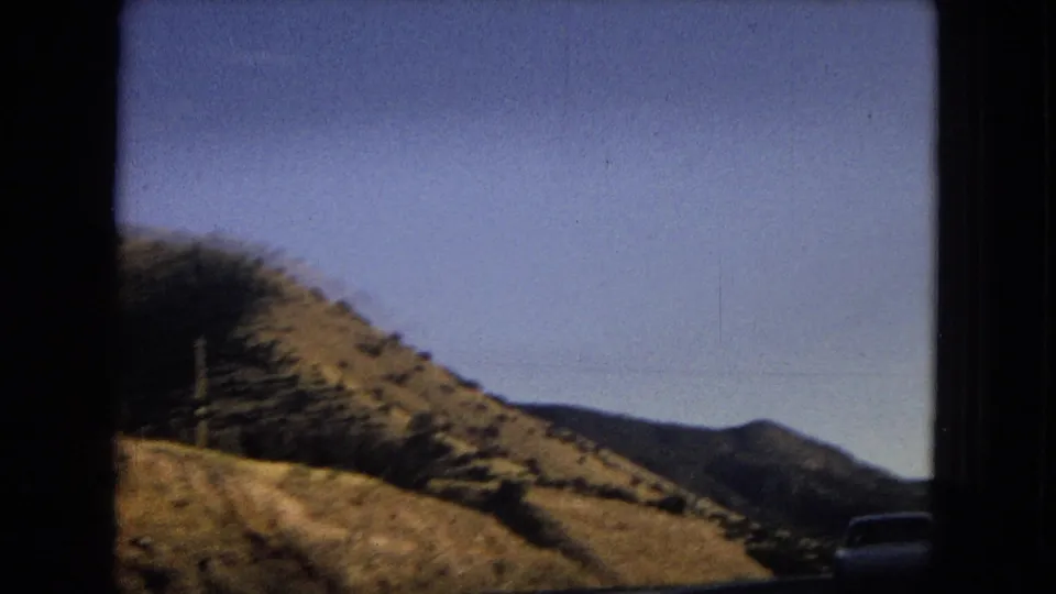 1967 Arizona road-trip frame showing dry hills, a road edge, blue sky, and a partially visible vehicle near the right side of the frame.