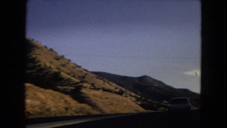 1967 8mm home-movie view from a moving car on a dry mountain road in the United States, with a white vehicle ahead and sunlit hillsides.