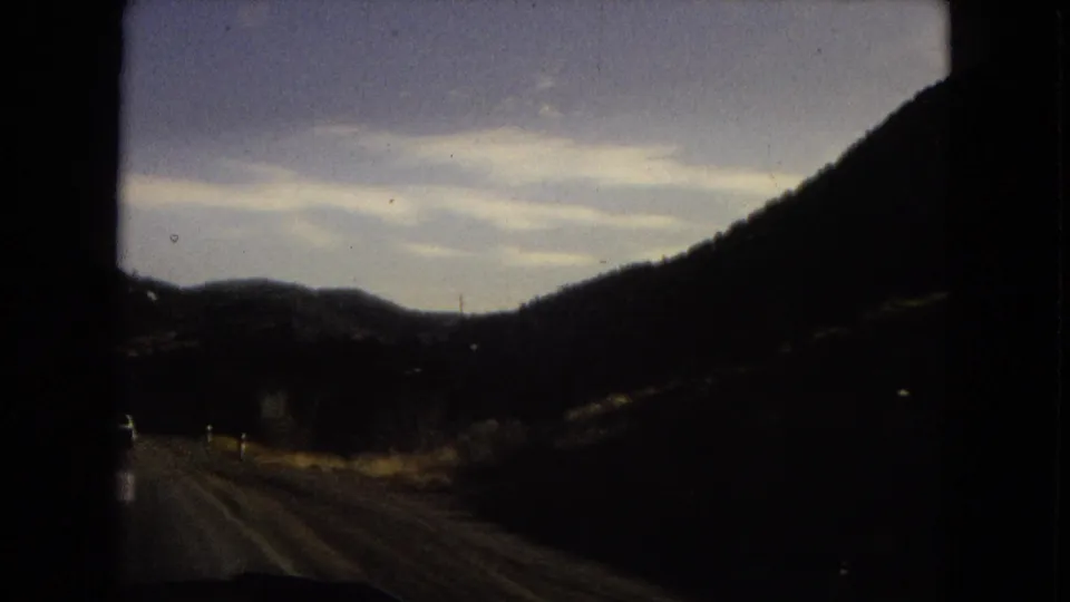 Passenger-perspective 8mm frame of a road passing between dark mountain slopes, with distant hills and a pale clouded sky.