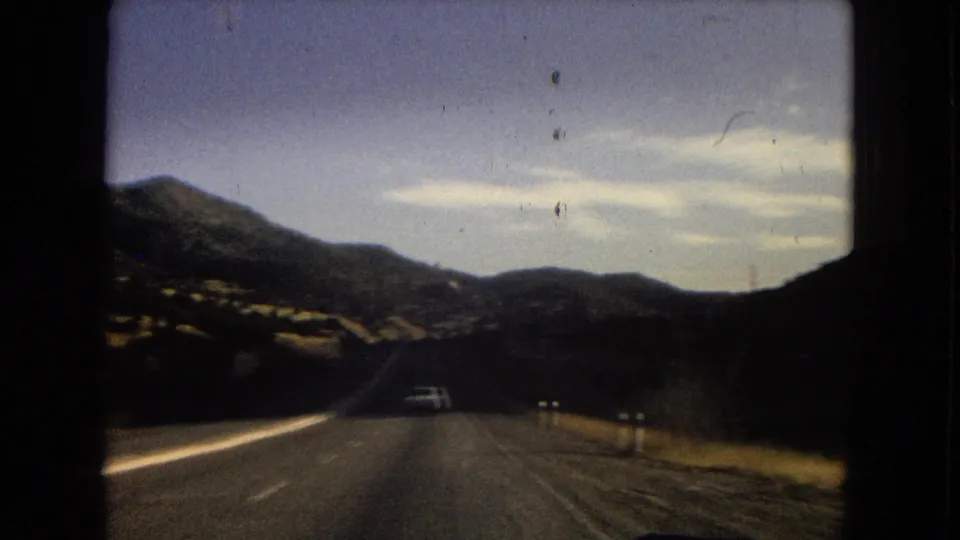 Vintage 8mm driving shot on a two-lane mountain highway, with dry slopes, distant vehicles, roadside posts, and open sky.