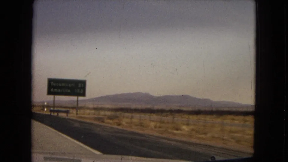 1967 8mm road footage showing an open highway across dry terrain, with a distance sign indicating Tucumcari and Amarillo against distant low mountains.