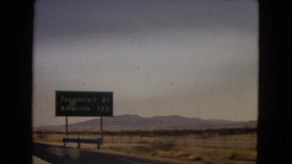 1967 8mm road-travel frame showing a highway sign for Tucumcari and Amarillo beside open fields and distant low mountains.