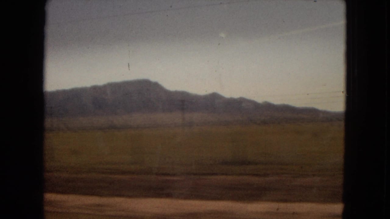 Open field and distant mountain range seen from a moving vehicle.