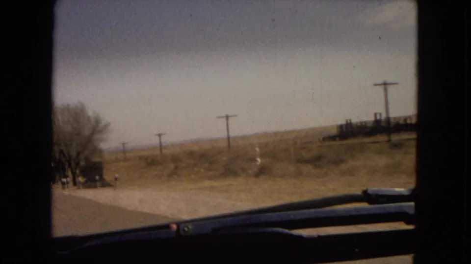 Moving-car view of a rural roadside with utility poles, sparse trees, fencing, a windshield wiper in the foreground, and small distant figures near the road.