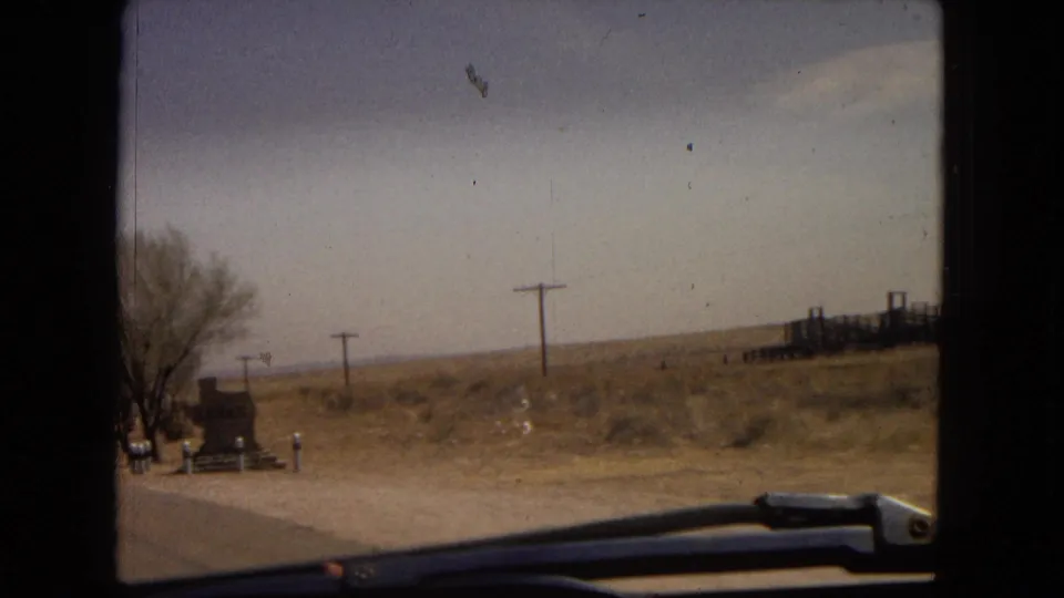 1967 8mm home-movie view from inside a moving car along a dry desert roadside in the American Southwest, with utility poles, sparse vegetation, and windshield hardware visible.