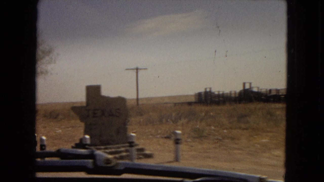 1967 8mm home-movie frame showing a Texas-shaped stone roadside marker viewed through a car windshield, with dry grass, utility poles, and low desert terrain beyond.