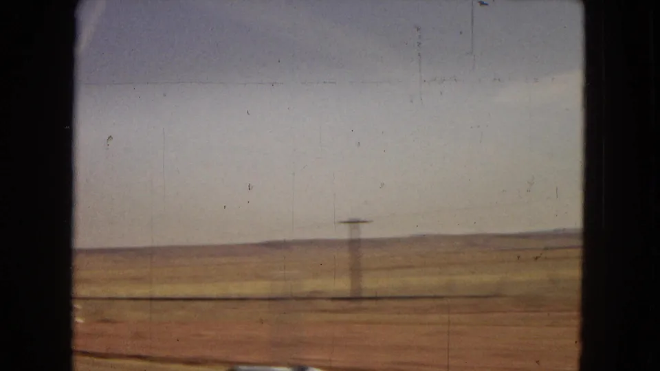 A moving 8mm car-window view of open Southwestern desert, showing a blurred utility pole, dry earth, low vegetation, and distant low hills under a pale sky.