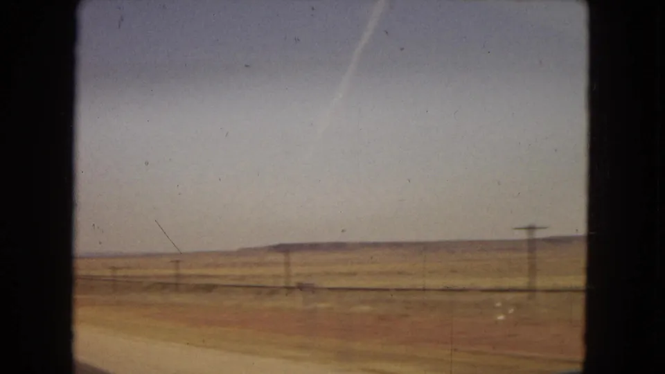 1967 8mm home-movie frame from a moving car across dry Southwestern terrain, with roadside earth, sparse brush, a utility pole, and low hills in the distance.