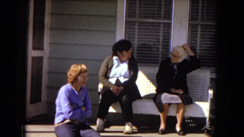 In 1967 Arizona home-movie footage, three women from different generations sit on a front porch, with one on the steps and two on a white bench beside shuttered windows.