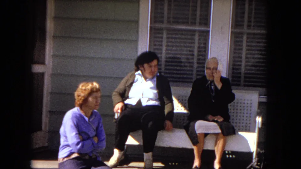 Three women sit in bright sunlight on the front porch, with the red-haired woman on the step, the center woman leaning back on the white bench, and the elderly woman seated beside her.