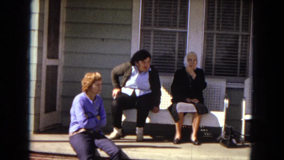 The final frame shows the three women still on the raised front porch in 1967 Arizona, framed by siding, windows, a door, and the white bench under strong sun.