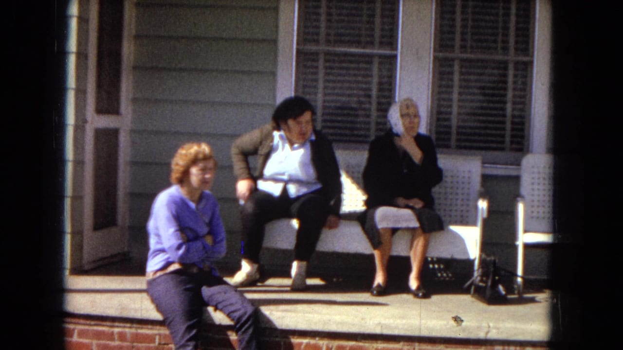 Three women sit casually on a porch in 1967 Arizona home-movie footage, with house siding, window shutters, and a white plastic chair visible.