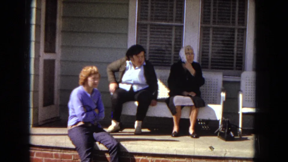 Three women sit casually on a porch in 1967 Arizona home-movie footage, with house siding, window shutters, and a white plastic chair visible.