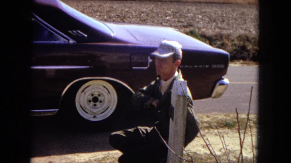A man in a checkered cap and dark jacket squats beside a purple 1960s car with visible rear wheel and model badging in 1967 Arizona footage.