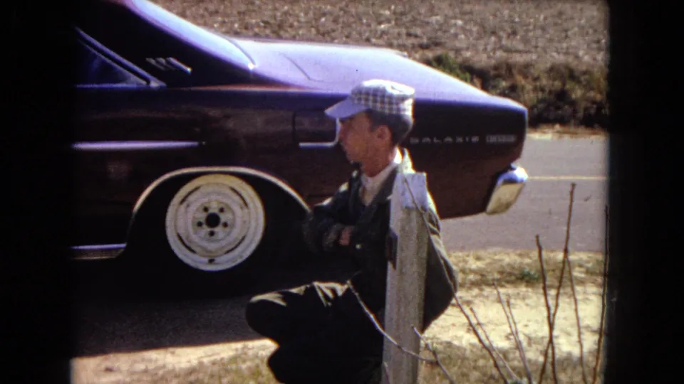 The man is shown in profile beside the purple car, with the rear quarter panel, white wheel, and dry roadside setting visible.