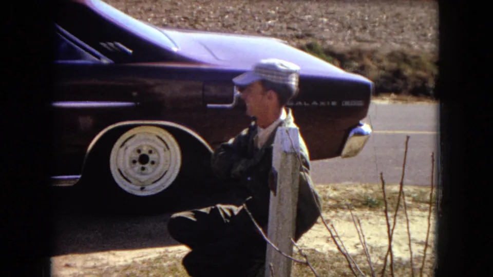A repeated close view shows the man squatting beside the purple car's rear wheel, with model badging, roadside pavement, and dry yard ground visible.
