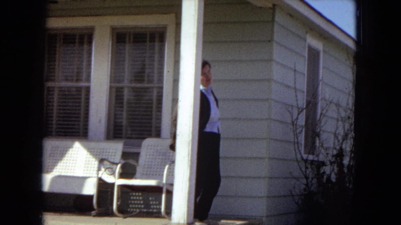 1967 United States 8mm home-movie frame showing a man in a dark business suit partly visible beside a modest farmhouse porch with white chairs and louvered windows.