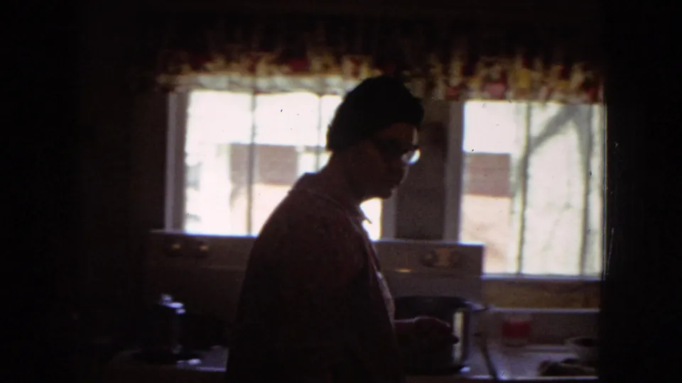 Dim 1967 home-movie kitchen frame showing a woman in a patterned dress and head covering working near bright windows, patterned curtains, cabinets, and a countertop.