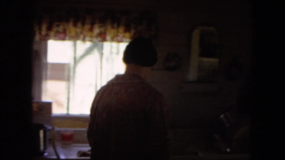 1967 United States 8mm frame showing a woman from behind in a dim farmhouse kitchen, standing near a counter below bright windows and patterned curtains.