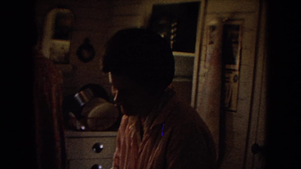 Shadowed 8mm frame of a private kitchen interior, with a person partly visible near cabinets and household objects in a 1967 Arizona home-movie context supplied by metadata.