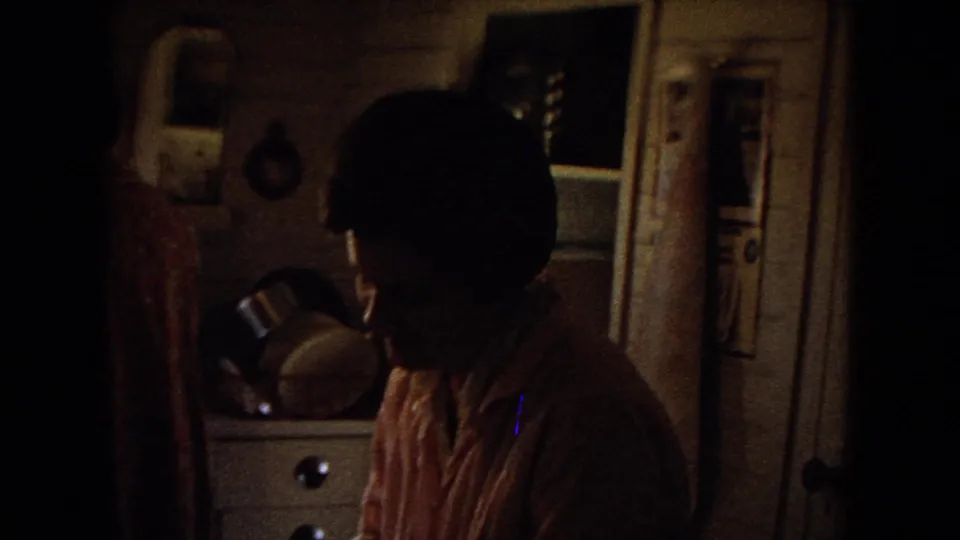 Shadowed 8mm frame of a private kitchen interior, with a person partly visible near cabinets and household objects in a 1967 Arizona home-movie context supplied by metadata.