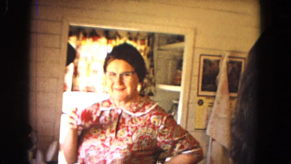 Woman in a floral dress and glasses smiles while holding a drink in a sunlit kitchen, consistent with the supplied 1967 Arizona home-movie metadata.