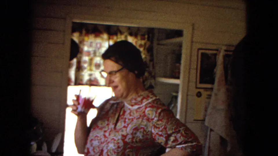 In a 1967 home-movie kitchen frame, the woman in a floral dress and glasses looks downward while holding a drink near patterned curtains and a door.