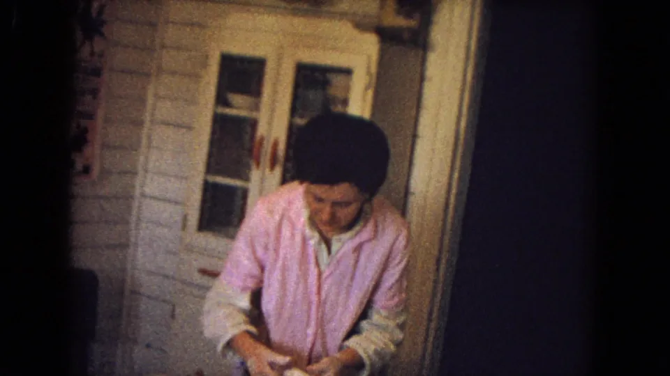 A person in pink clothing stands near a kitchen door and looks down, with paneled walls and household fixtures visible in the private interior.