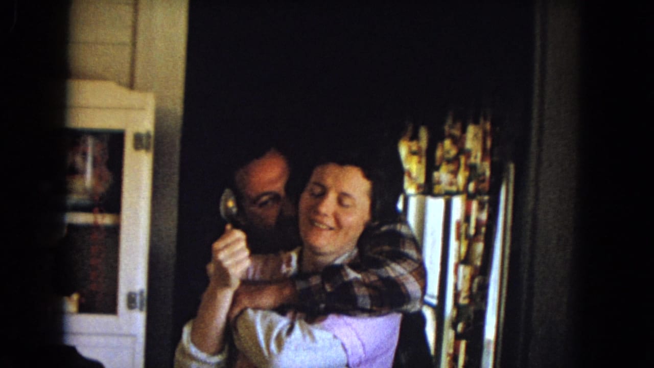 In 1960s United States 8mm home-movie footage, a man embraces a smiling woman inside a dim kitchen with floral curtains and cabinets nearby.