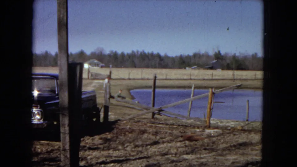 A rural United States property view shows a pond, rough wooden fencing, muddy ground, a dark truck, and distant low buildings or trees under a blue sky.