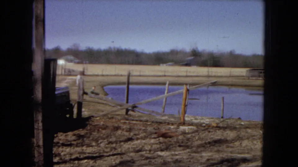The exterior view continues across the rural property, with a pond behind a rough fence, the edge of a dark truck, open muddy ground, and a distant tree line.
