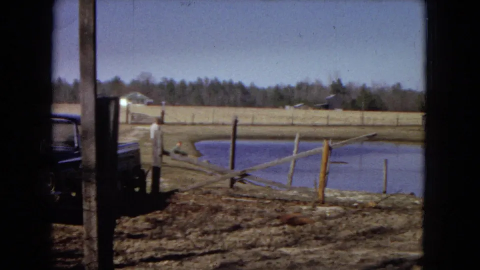 1967 home-movie frame of a small rural pond with fence posts, dry grass, a distant field, and a dark vehicle partly visible at the left edge.