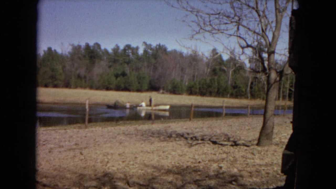 A small boat with people fishing sits on a rural pond, with fence posts, dry banks, bare trees, and woods in the background.