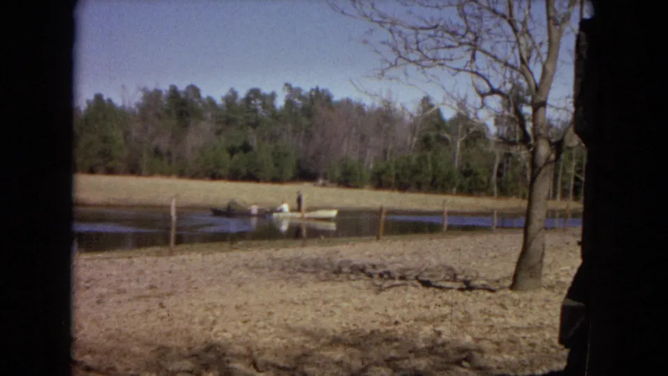 A small boat with people fishing sits on a rural pond, with fence posts, dry banks, bare trees, and woods in the background.