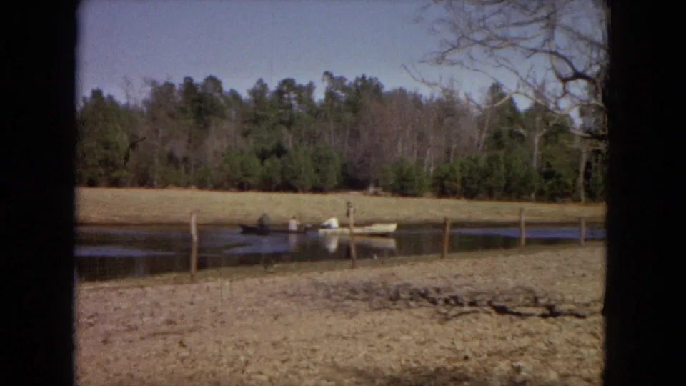 Home-movie view across a fenced rural pond toward a small boat with people fishing near a grassy bank and wooded tree line.