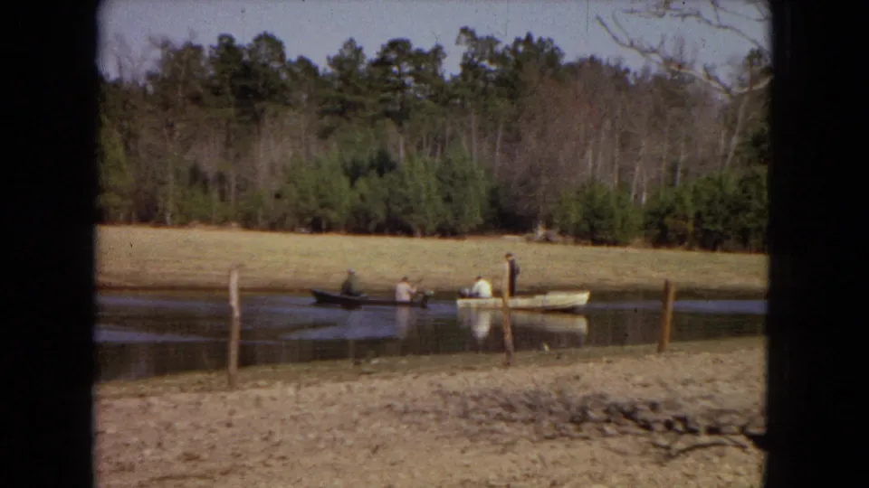 A small rowboat with people fishing rests near the center of a rural pond, framed by fence posts, dry grass, and a wooded background.