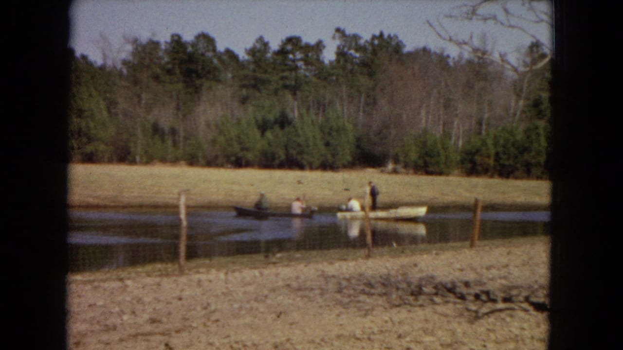 1967 home-movie frame showing two small boats with people on a calm pond bordered by dry grass and pine woods.
