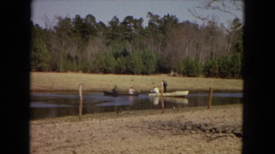 1967 home-movie frame showing two small boats with people on a calm pond bordered by dry grass and pine woods.
