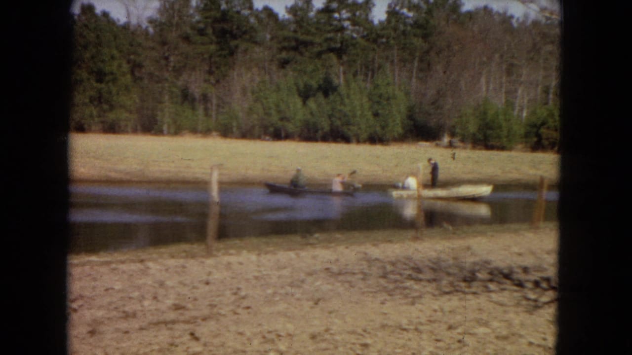 1967 home-movie view of people in two small boats crossing a pond near a grassy field and dense trees.