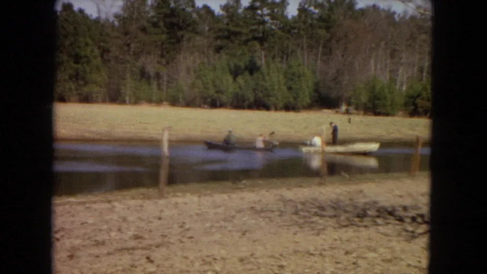 1967 home-movie view of people in two small boats crossing a pond near a grassy field and dense trees.
