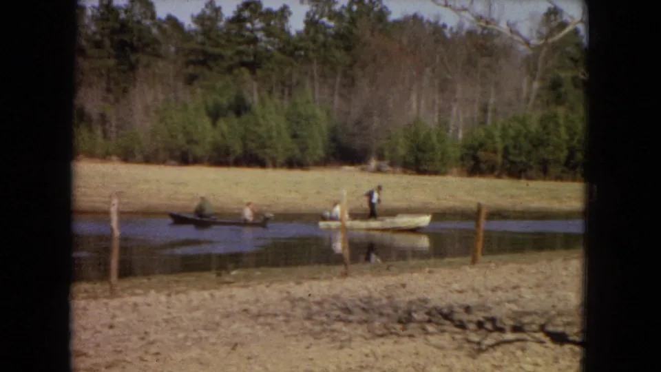 1967 8mm frame showing people in small boats on a pond, viewed past a dark foreground divider with woods in the background.