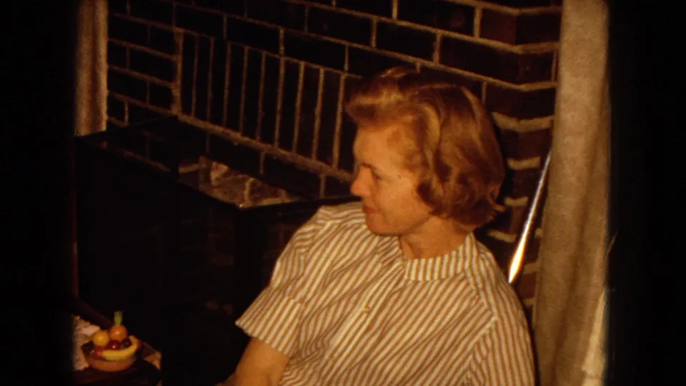 1967 home-movie frame of a woman in a striped blouse seated beside a brick fireplace in a private interior.