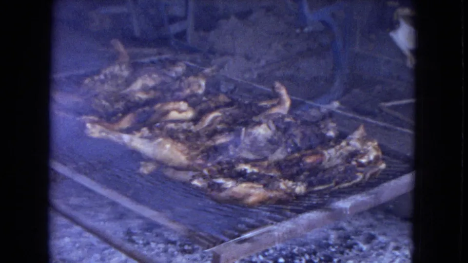 1967 home-movie frame showing whole chickens or large poultry pieces cooking on an outdoor metal grill, with smoke and a blue-tinted 8mm film look.